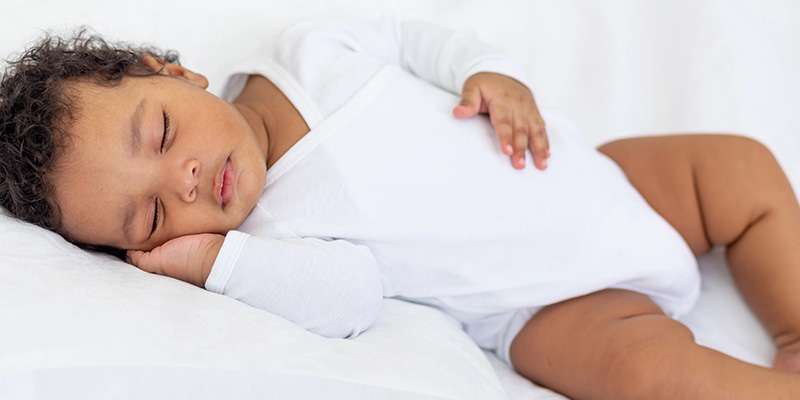 CDPH_FI-Safe Kids-Safe Sleep Cobb & Douglas Public Health: A baby wearing a white onesie sleeps peacefully on its side on a white bed, with one hand resting on its stomach and the other under its head.